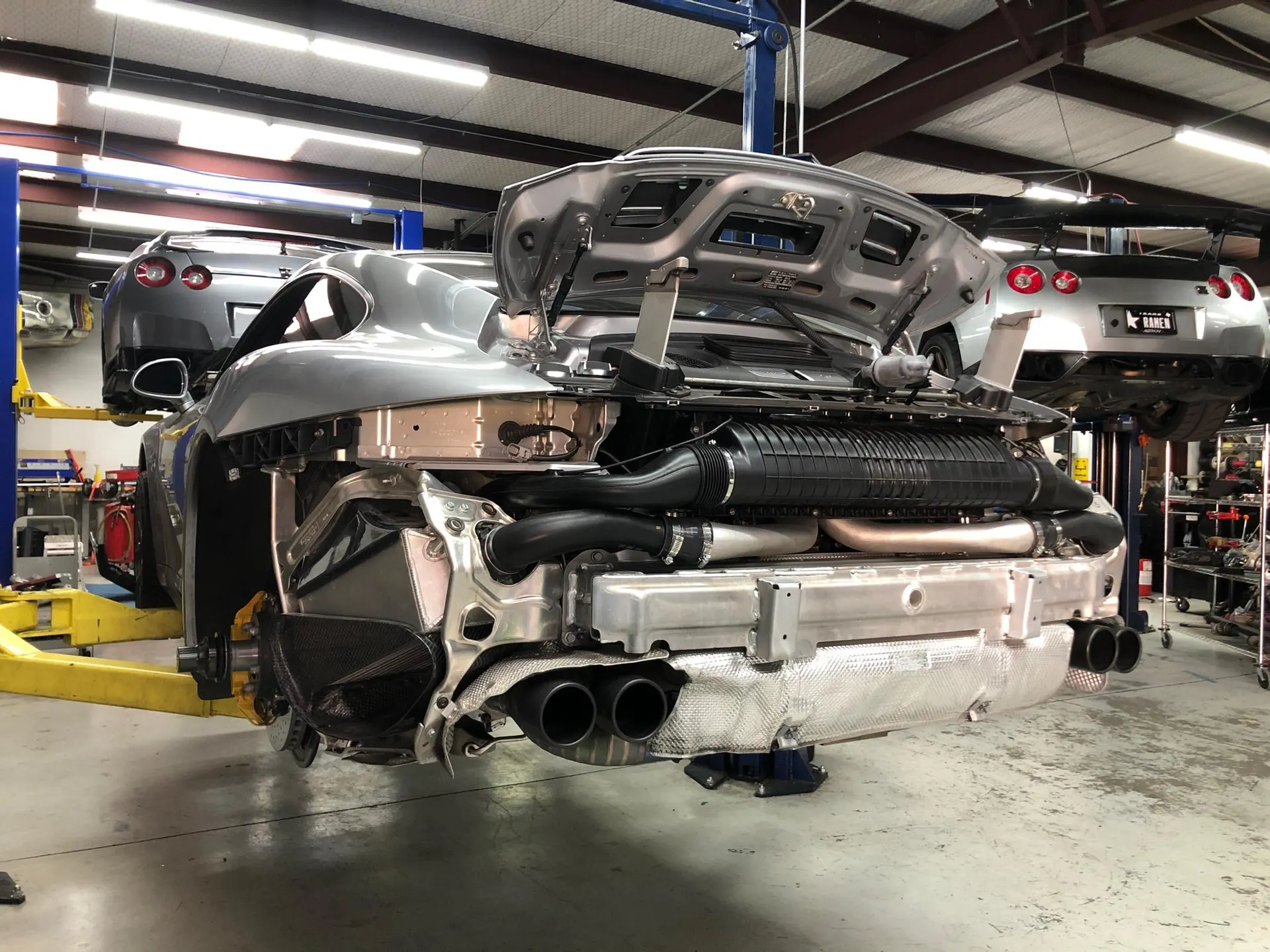 A silver Porsche 911 on a lift in a performance shop with its rear bumper removed, exposing the exhaust and engine components.