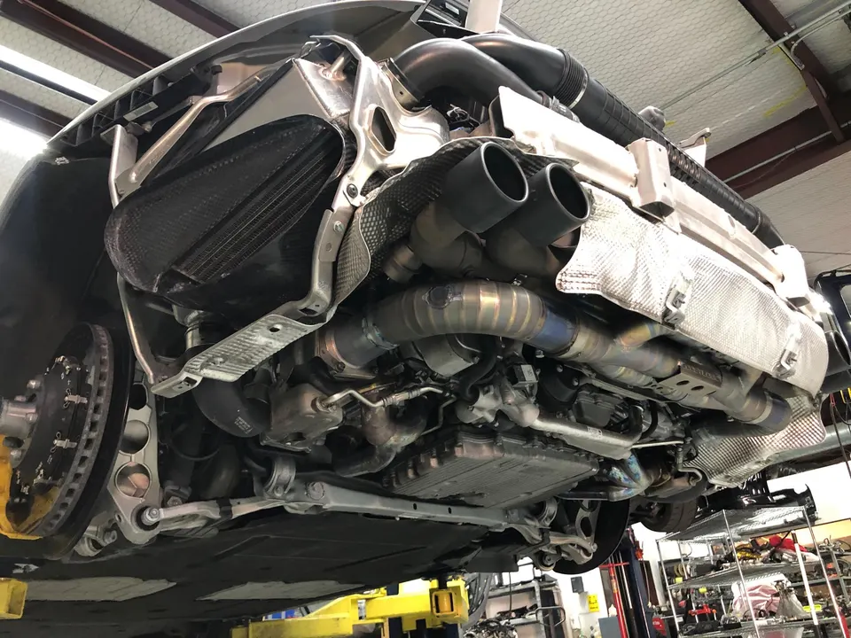 Underside view of a Porsche on a lift in a shop, showcasing a titanium exhaust system and carbon fiber components.