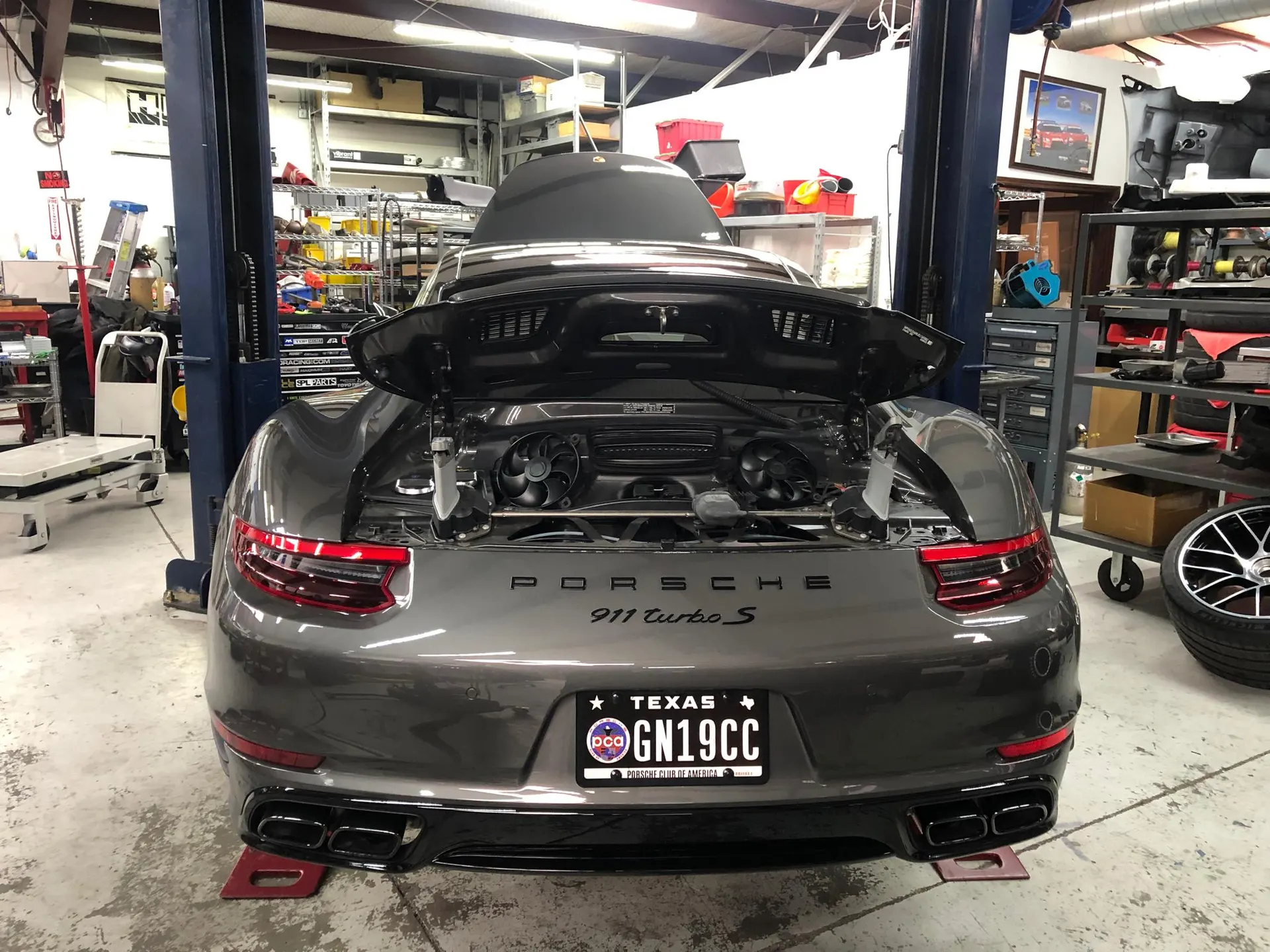 A grey Porsche 911 Turbo S on a lift in a shop with its engine bay open, showing the rear engine and exhaust.