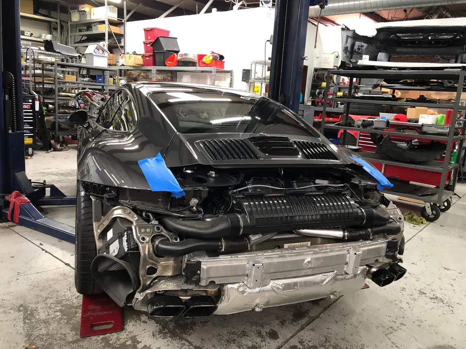 A grey Porsche 911 in a shop with its rear bumper removed, exposing the induction system and exhaust tips.