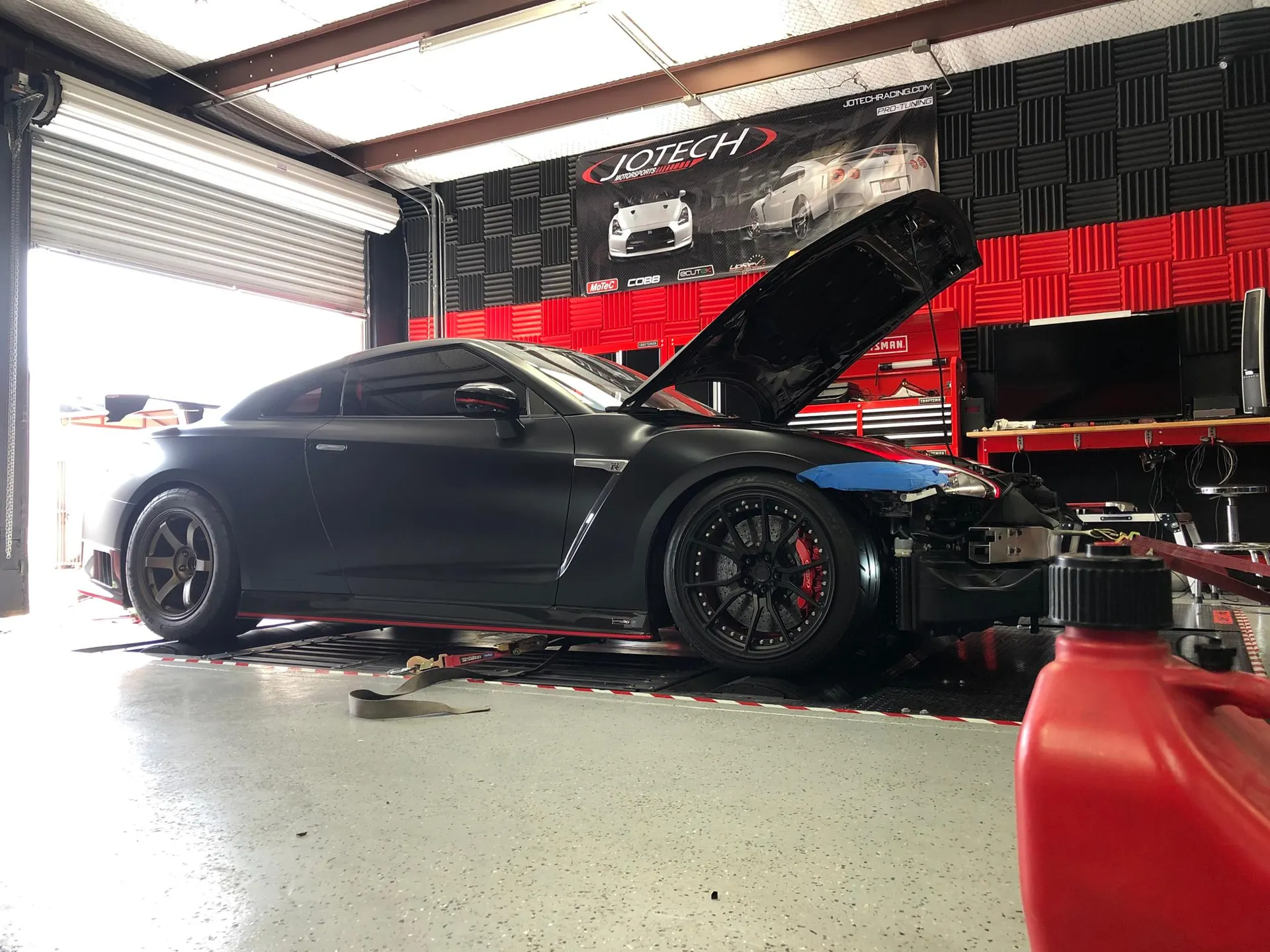 A matte black Nissan GT-R on a dyno at Jotech Motorsports with the hood open and front bumper removed for tuning.