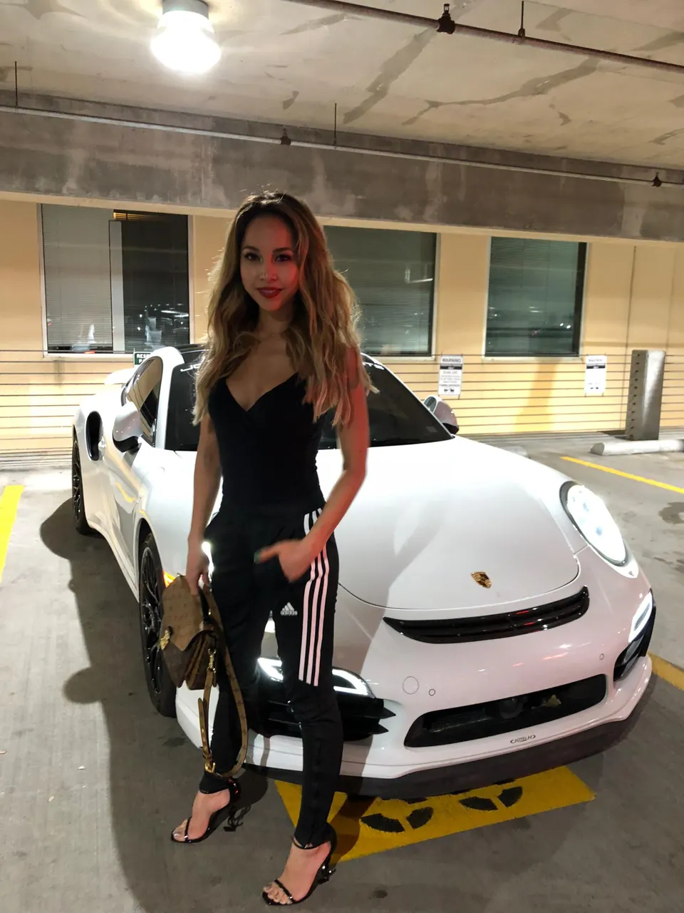 A woman in black athletic wear and heels stands next to a white Porsche sports car in a parking garage.