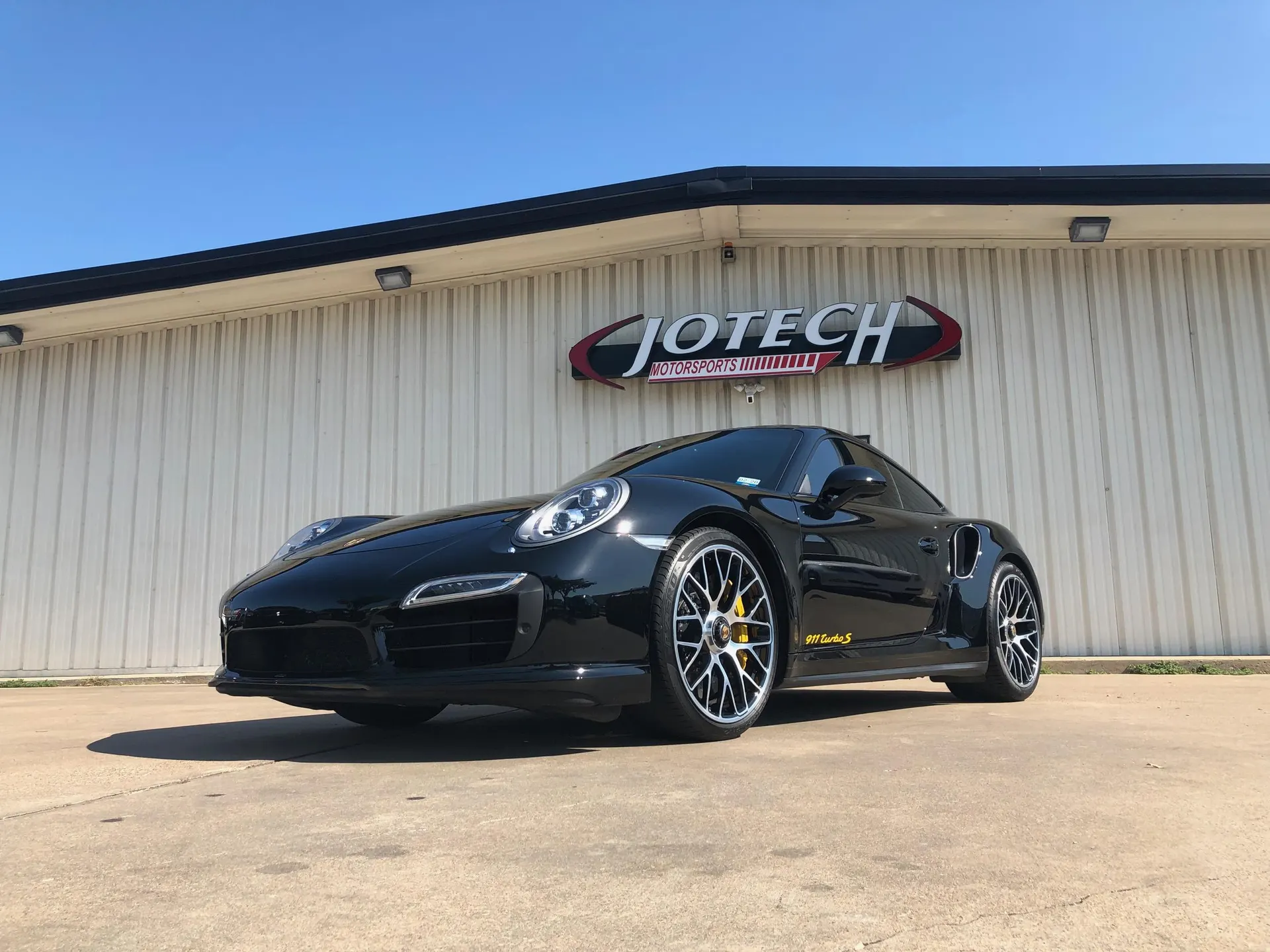Black Porsche 911 Turbo S parked outside Jotech Motorsports, showcasing its sleek design and performance wheels.
