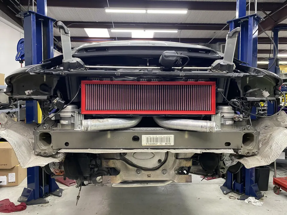 A Porsche 911 on a lift in a shop, with its rear bumper removed, showcasing a large K&N air filter and boost piping.
