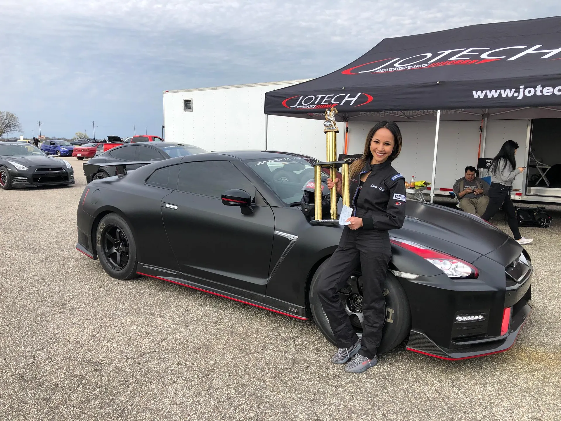 Woman in racing suit holding a trophy next to a matte black Nissan GT-R at a Jotech Motorsports track event.