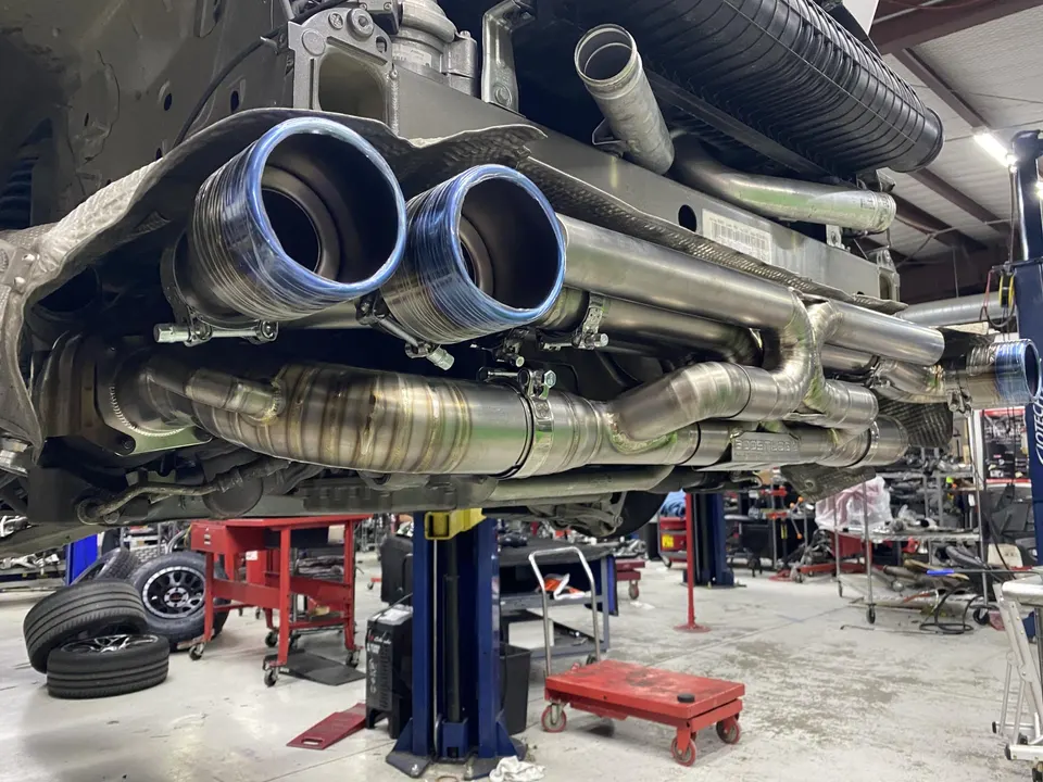 Custom titanium exhaust system with blue-tipped mufflers installed on the underside of a car on a lift in a performance shop.