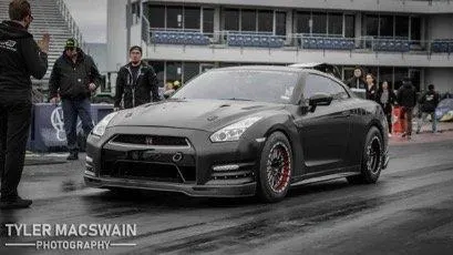 Black Nissan GT-R with aftermarket wheels in the staging lane of a drag strip, with people and bleachers in the background.