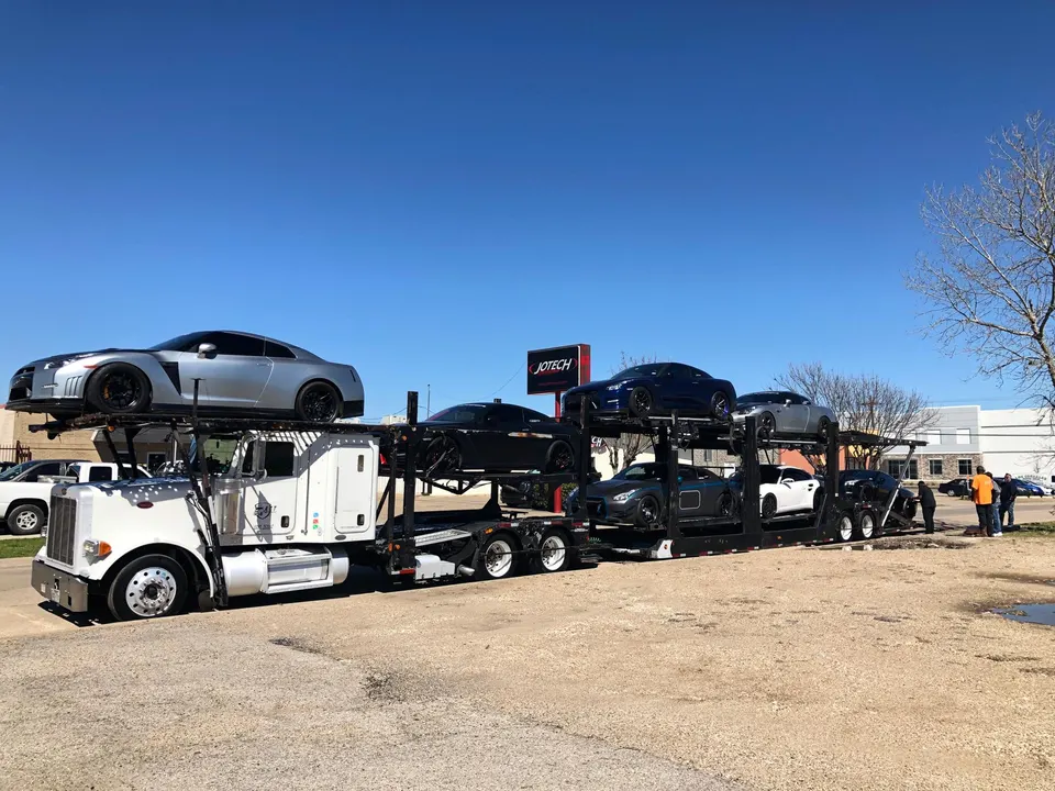 A car hauler truck loaded with multiple high-performance sports cars, including Nissan GT-Rs and a Mercedes-AMG GT, parked outside Jotech Motorsports.