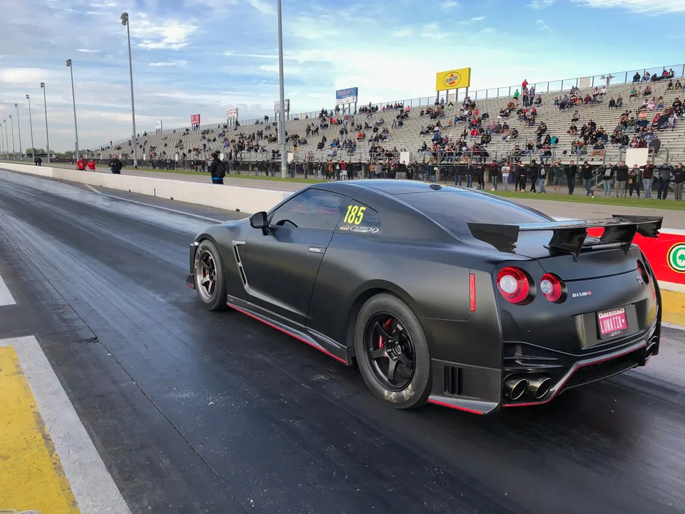 Matte black Nissan GT-R race car with a large rear wing on a drag strip, with spectators in the stands.