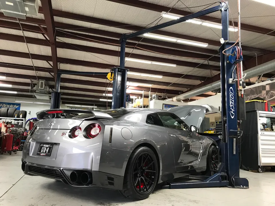 A silver Nissan GT-R on a car lift in a performance tuning shop with its hood open, viewed from the rear quarter.