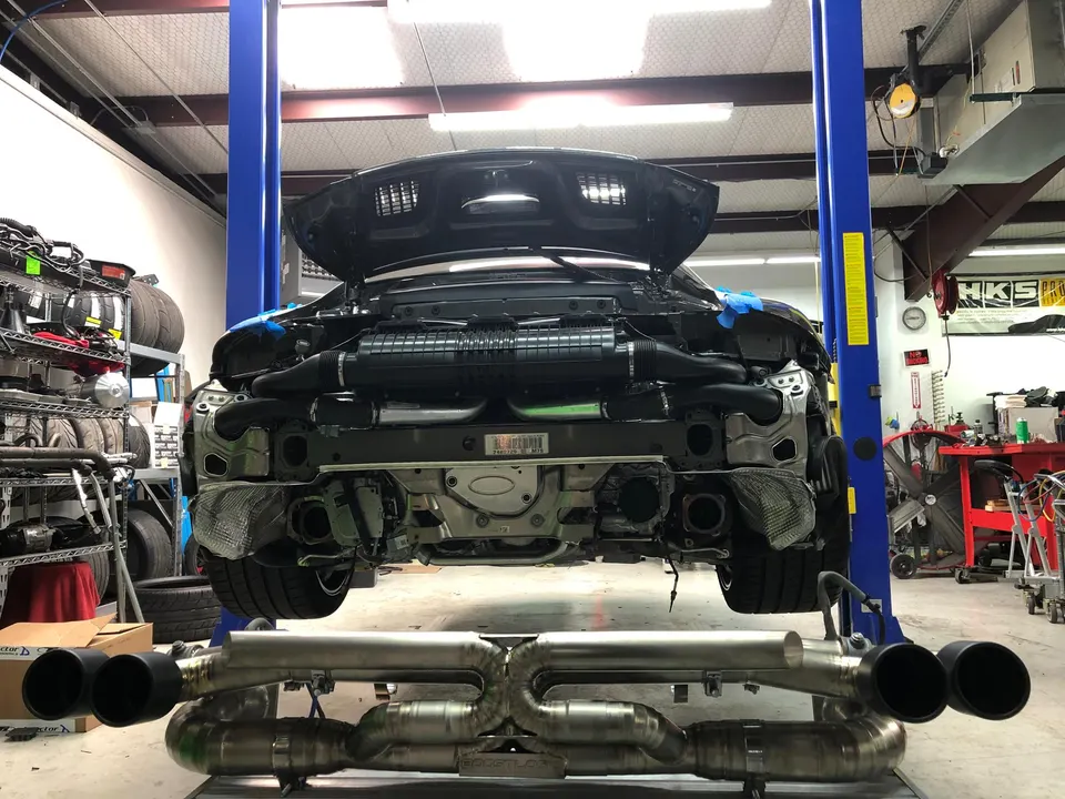 Porsche 911 on a lift in a performance shop with a new titanium exhaust system ready for installation.