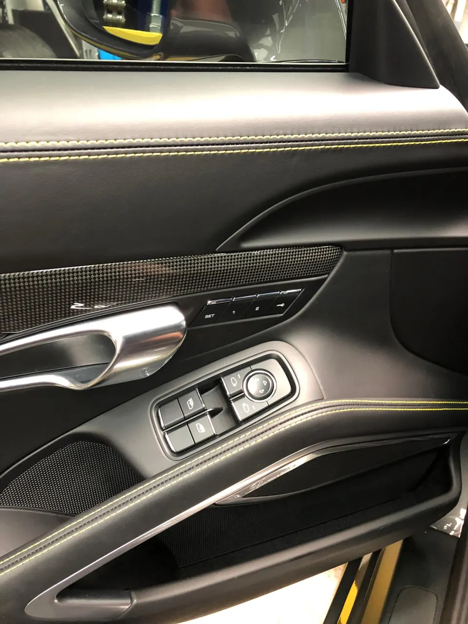 Close-up of a luxury car's interior door panel featuring black leather with yellow stitching, carbon fiber trim, and window controls.