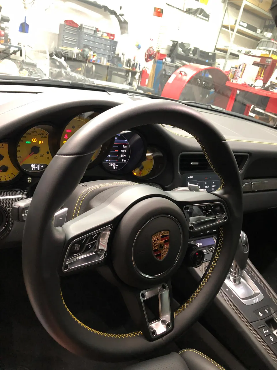 A Porsche interior featuring a black steering wheel with yellow stitching and a dashboard, seen inside a car shop.