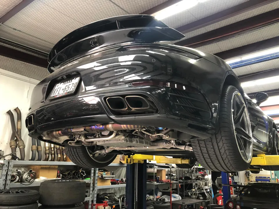 A black Porsche 911 on a car lift in a performance shop, showcasing its aftermarket titanium exhaust system.