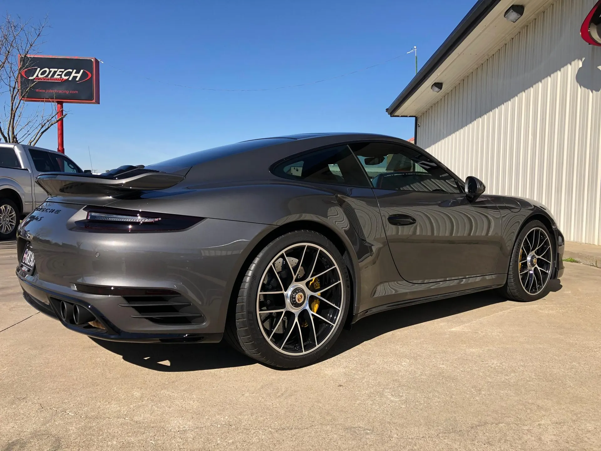 A grey Porsche 911 Turbo S parked outside Jotech Racing under a clear blue sky.