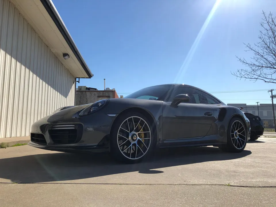 Grey Porsche 911 sports car parked outdoors in a sunny parking lot next to a building.