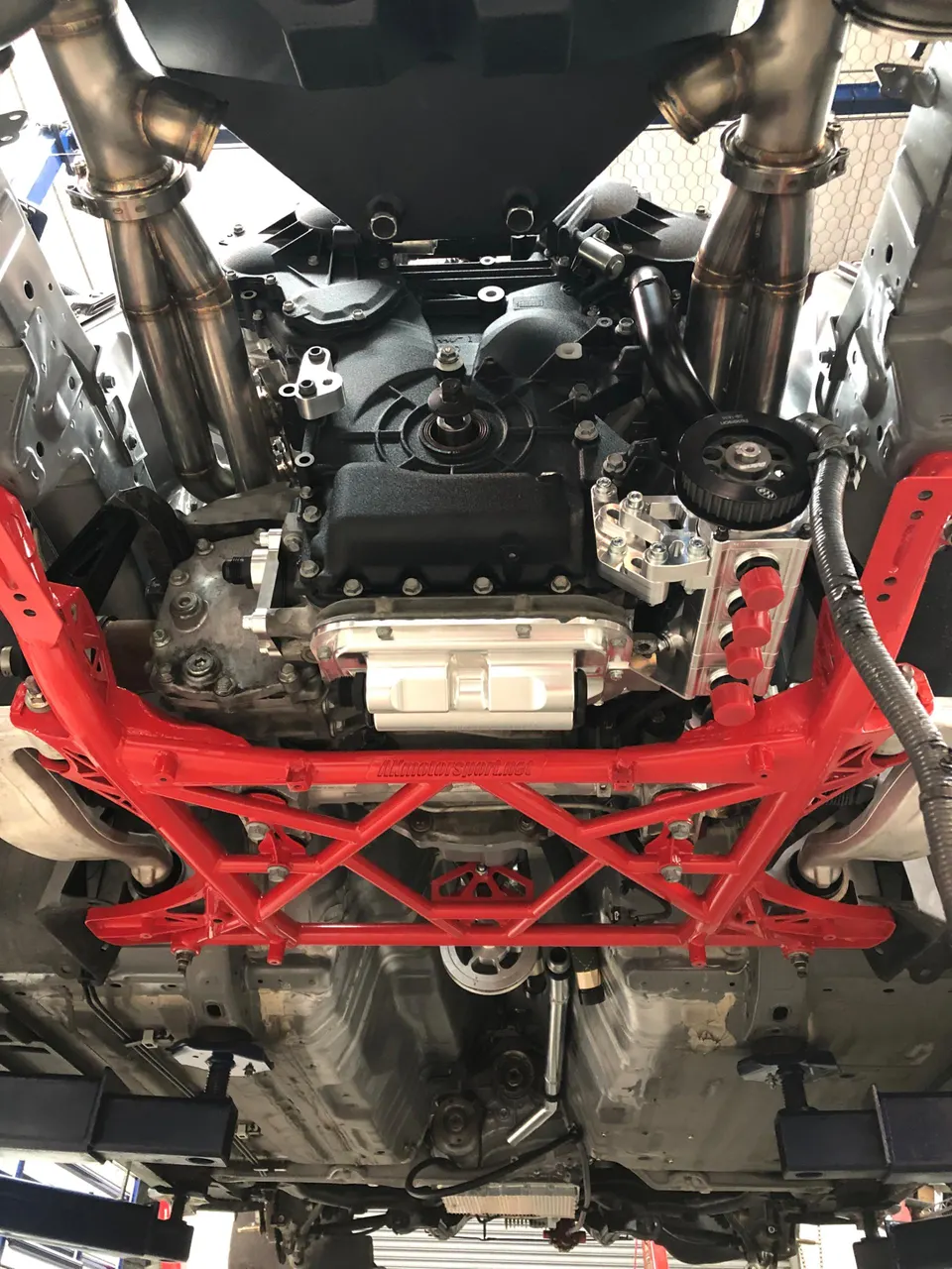 Underside view of a performance car on a lift, showcasing a red aftermarket chassis brace, custom exhaust headers, and engine.