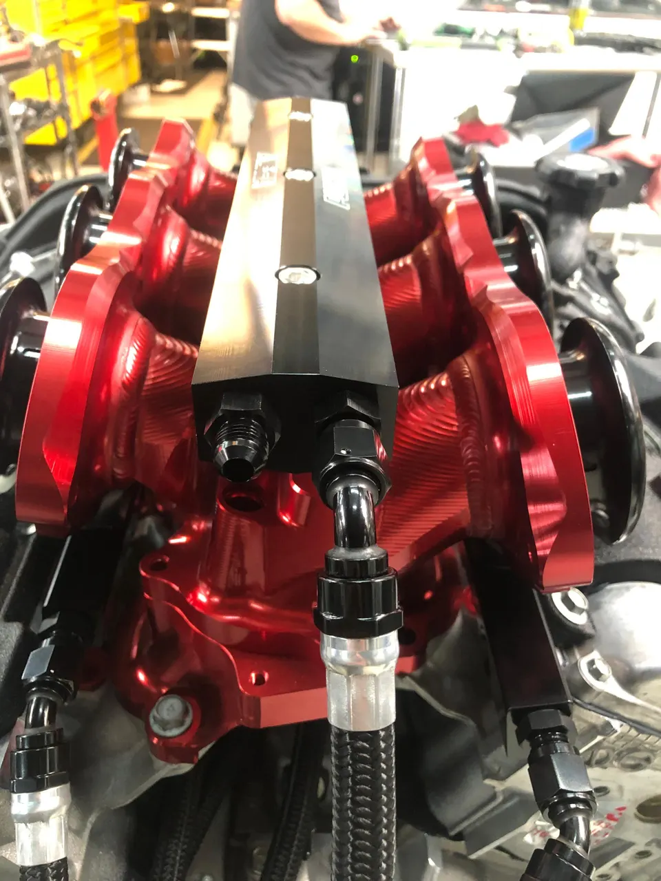 Close-up of a red aftermarket intake manifold with black velocity stacks and braided fuel lines being installed in a shop.