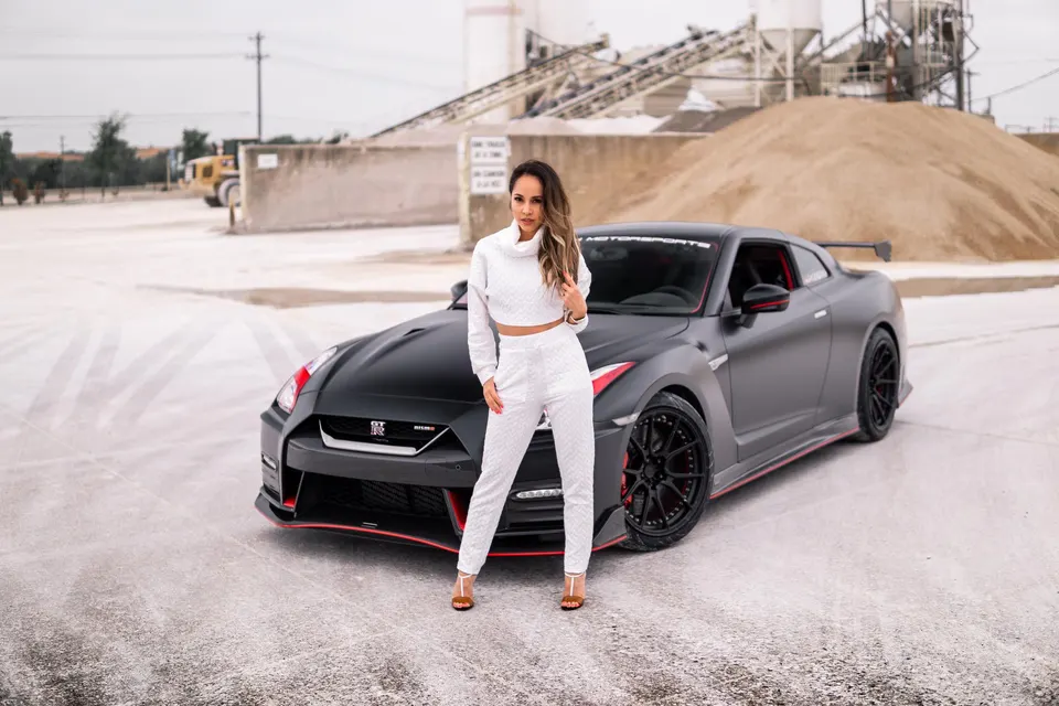 A woman in white posing next to a matte black Nissan GT-R with red accents and aftermarket wheels at an industrial site.