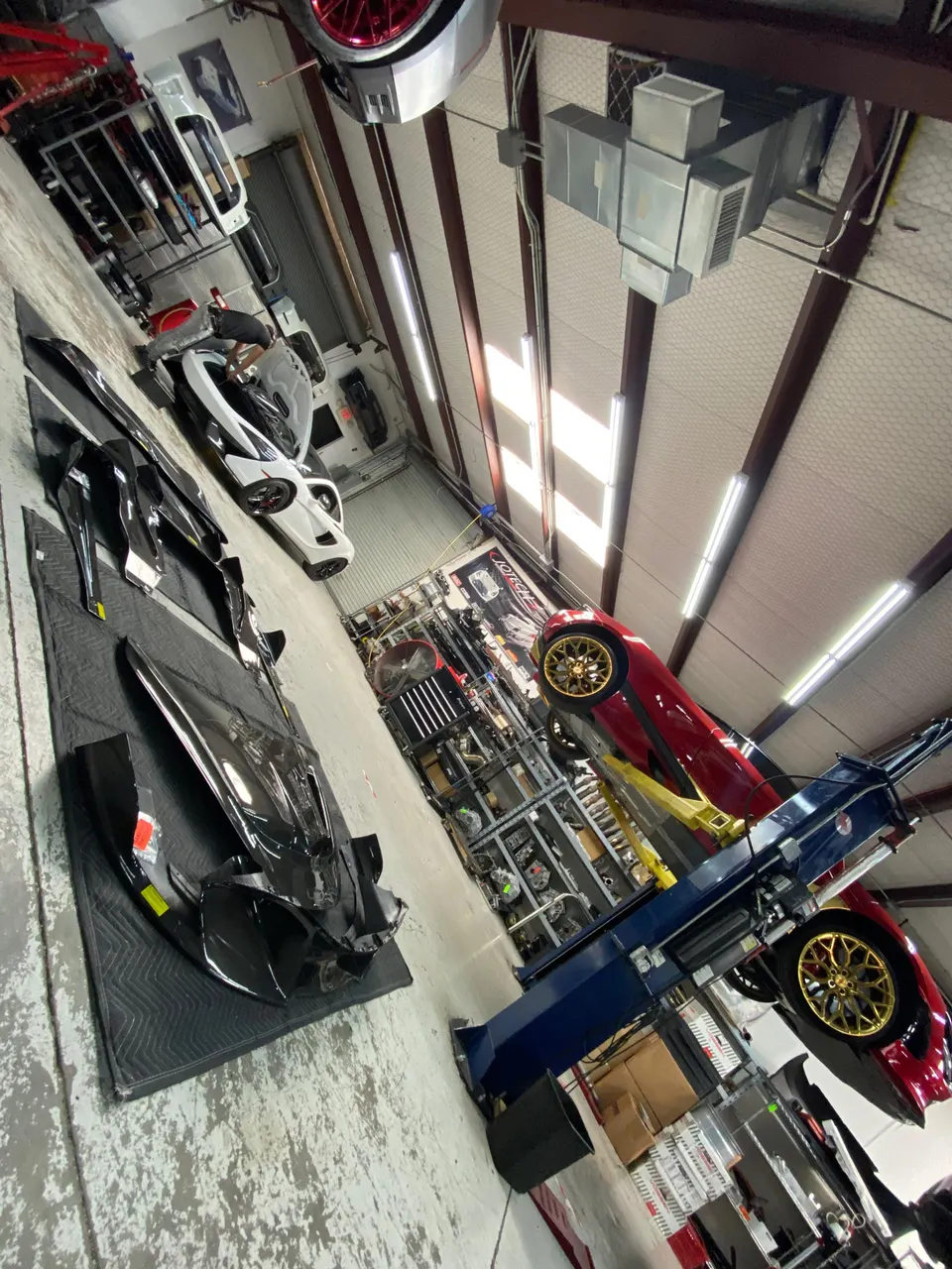 A busy performance shop interior with a white McLaren and a red Toyota Supra on a lift, surrounded by car parts.
