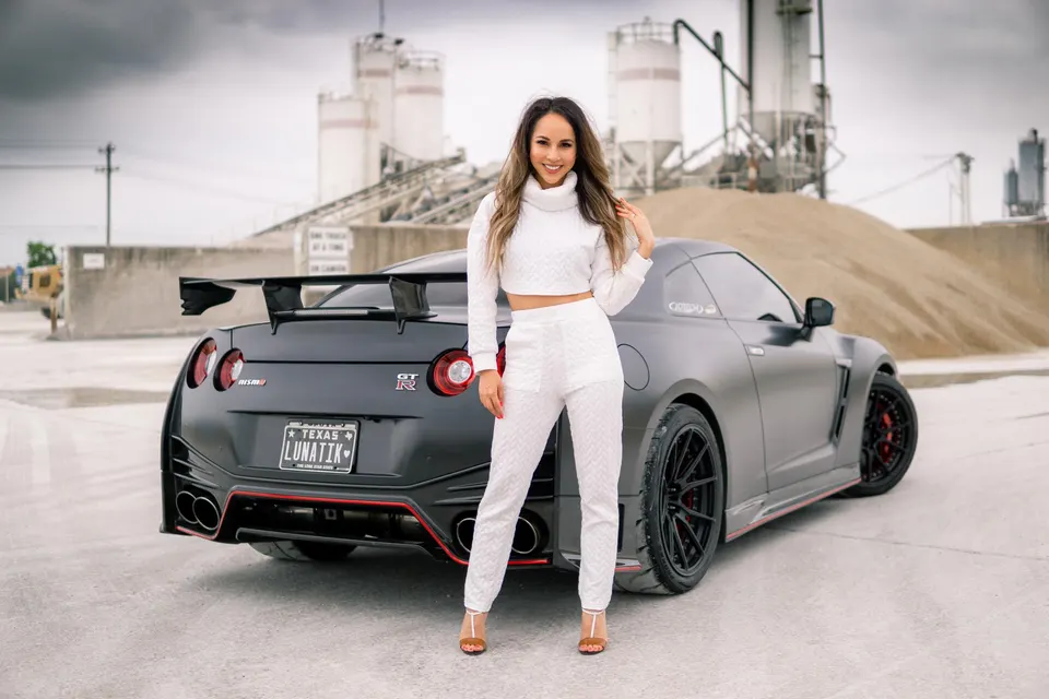 A woman in white posing next to a matte black Nissan GT-R with a large rear wing and aftermarket wheels at an industrial site.