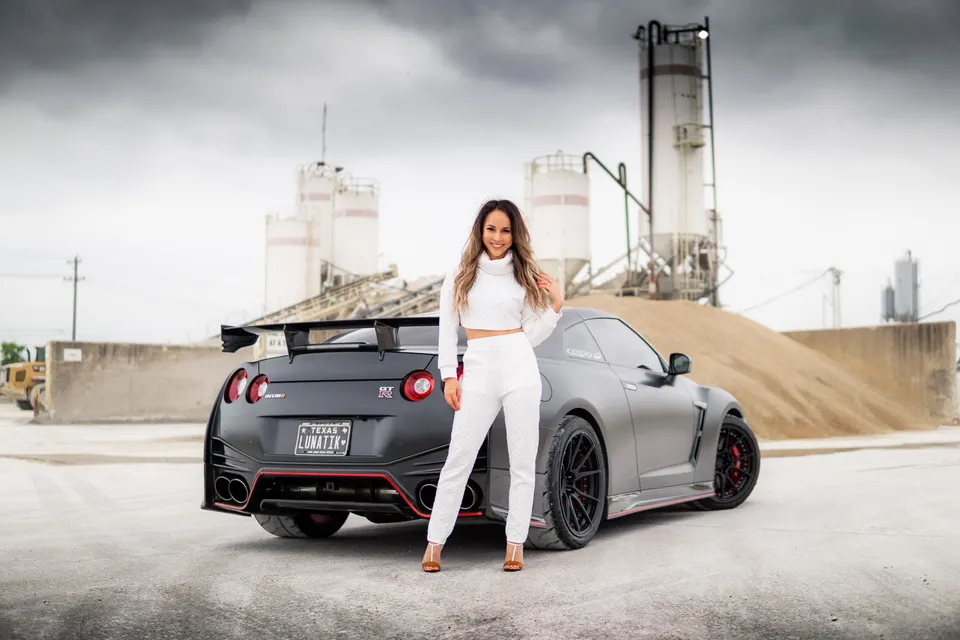 Matte black Nissan GT-R R35 with a large rear wing and aftermarket wheels, parked in an industrial setting with a woman.
