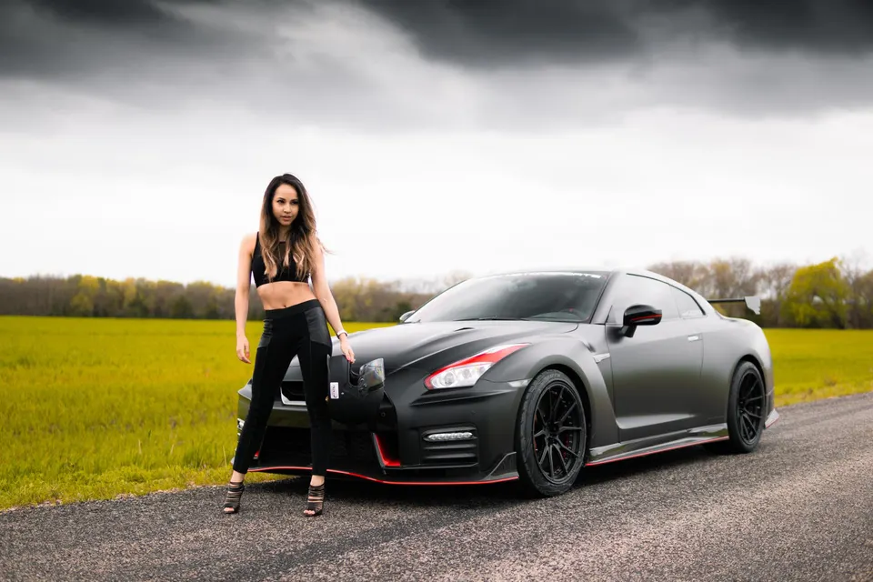 Matte black Nissan GT-R with red accents parked on an asphalt road with a woman posing next to it in a field.