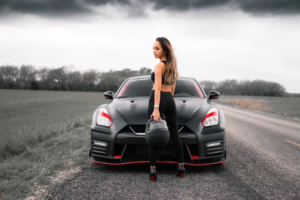 A woman holding a helmet stands in front of a black Nissan GT-R on a rural road under a cloudy sky.