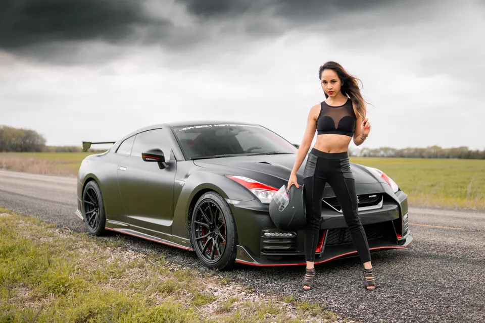 Matte grey Nissan GT-R with red accents and aftermarket wing, parked on a road with a model holding a helmet.