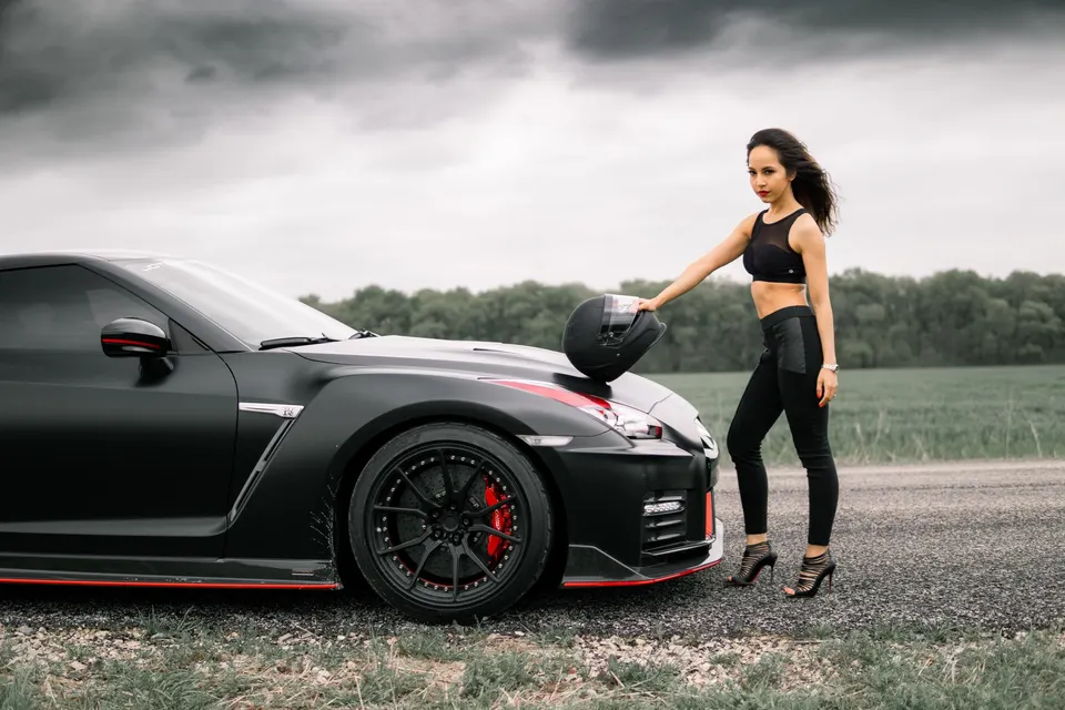 A woman in black attire holding a helmet next to a matte black Nissan GT-R with custom wheels and red accents.