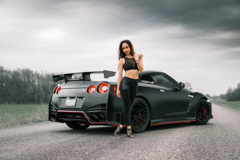 A woman stands next to a matte black Nissan GT-R with a large aftermarket wing and custom wheels on an overcast day.