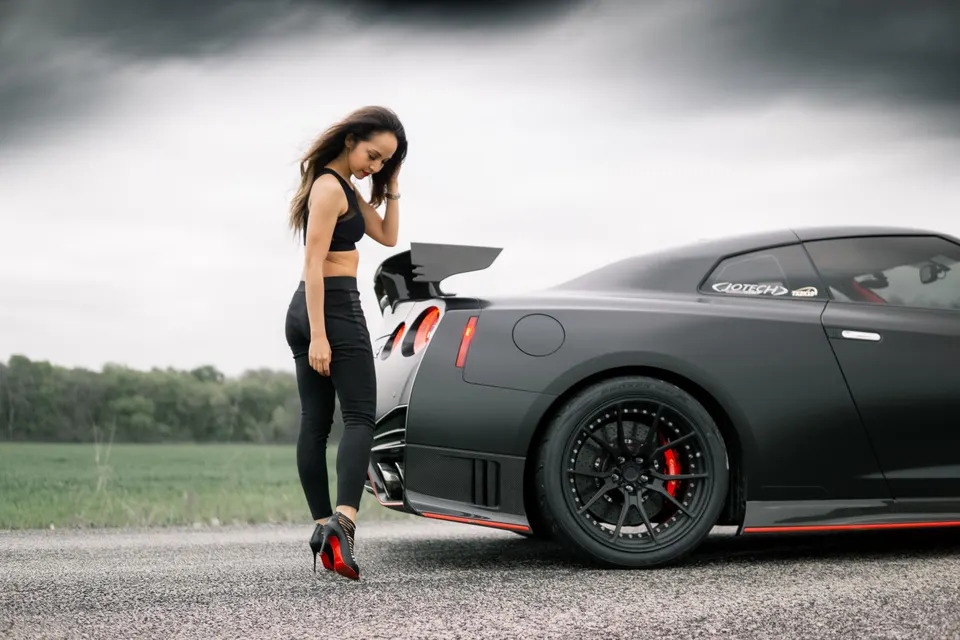 Matte black Nissan GT-R with an aftermarket wing and custom wheels, parked outdoors with a woman standing beside it.