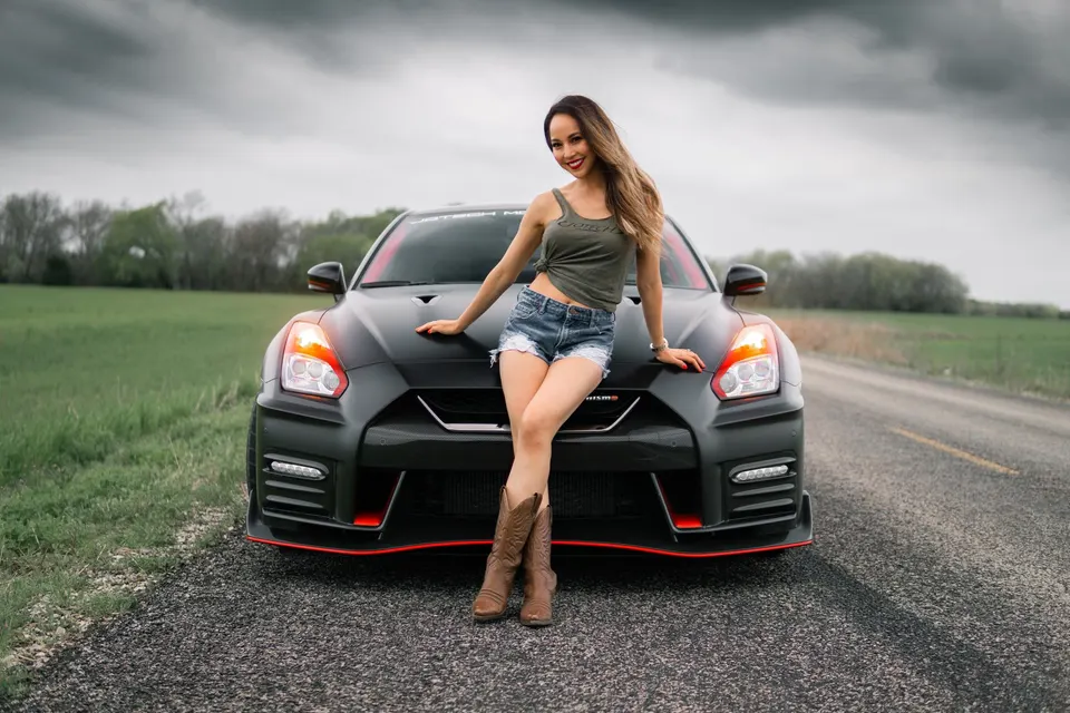 A woman in denim shorts and a tank top poses on the hood of a black Nissan GT-R on a rural road under a cloudy sky.