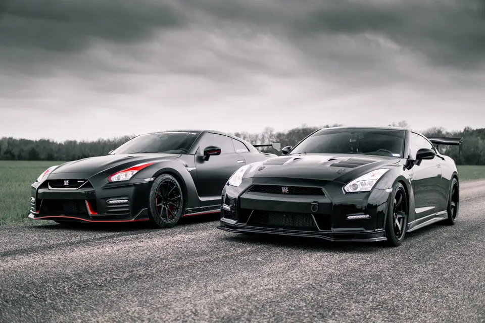 Two black modified Nissan GT-R R35 sports cars parked on an asphalt road under a cloudy sky.