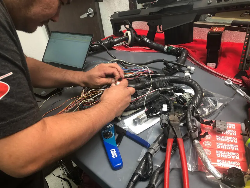 Technician working on a complex wiring harness with a laptop and tools in a performance tuning shop.