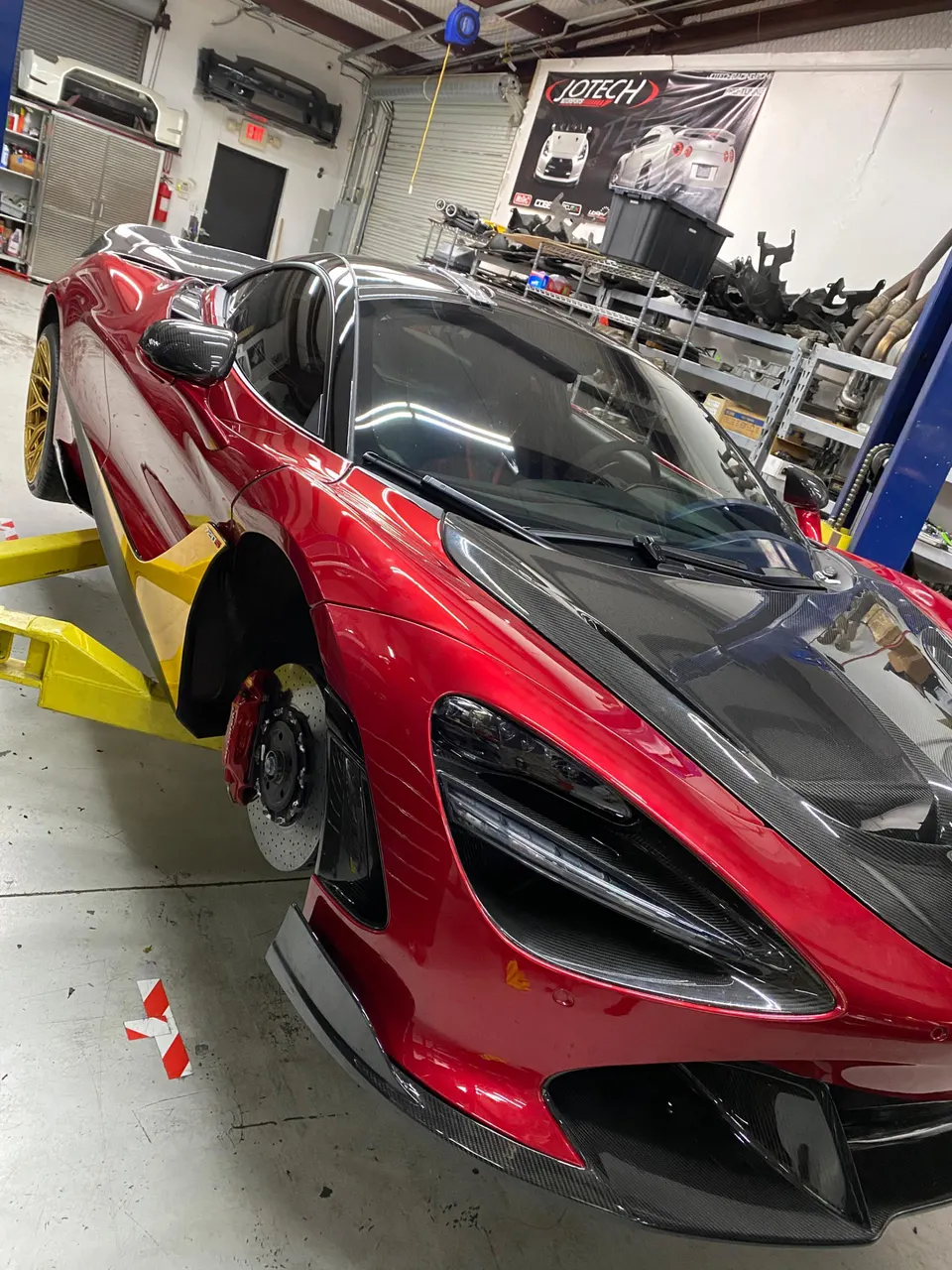 A red McLaren 720S on a lift in a performance tuning shop, featuring carbon fiber body parts and visible brakes.