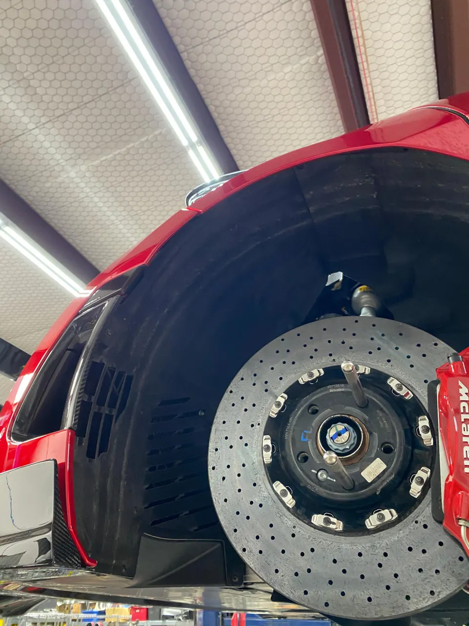 Close-up of a red McLaren's carbon ceramic brake rotor and red caliper, with suspension components visible in a shop.