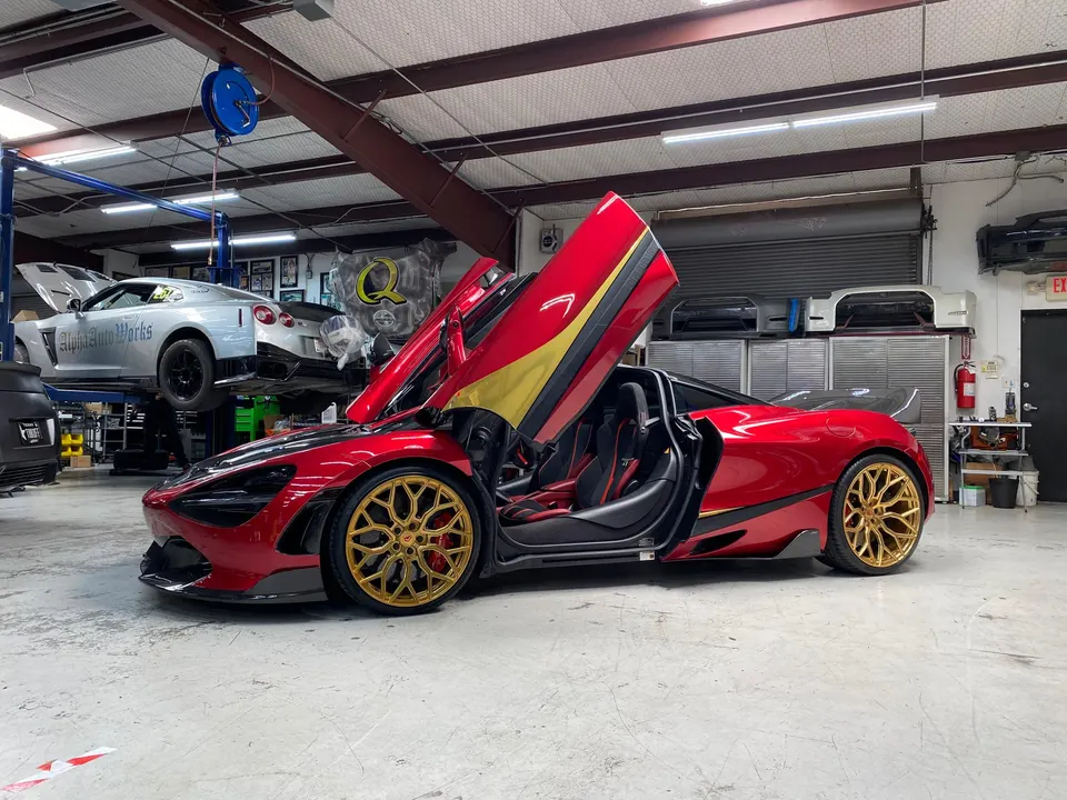 A red McLaren 720S with gold wheels and open doors inside a car performance shop, with a silver Nissan GT-R on a lift.