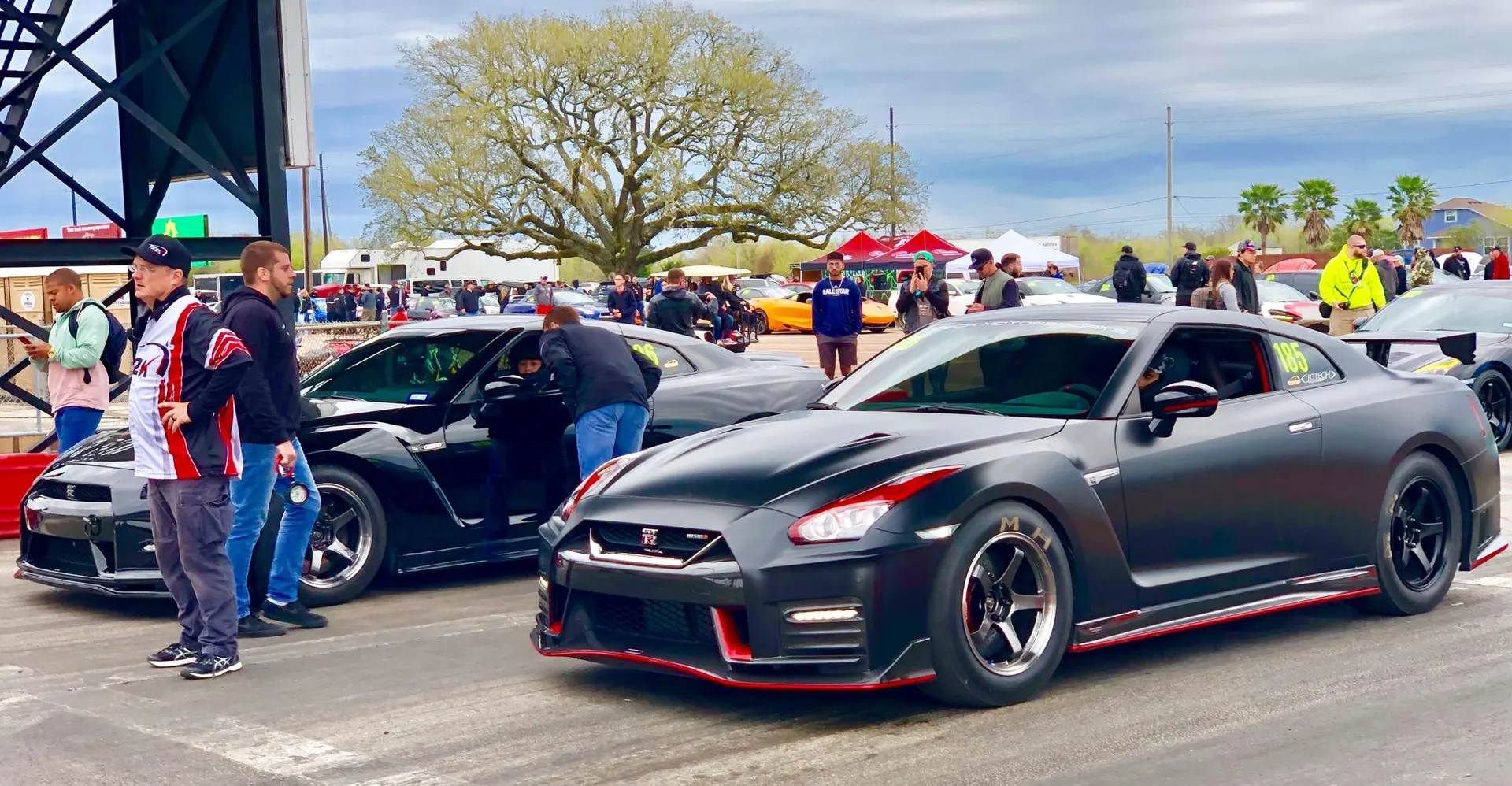 Two modified Nissan GT-Rs, one matte black with red accents, at a drag strip event with spectators.