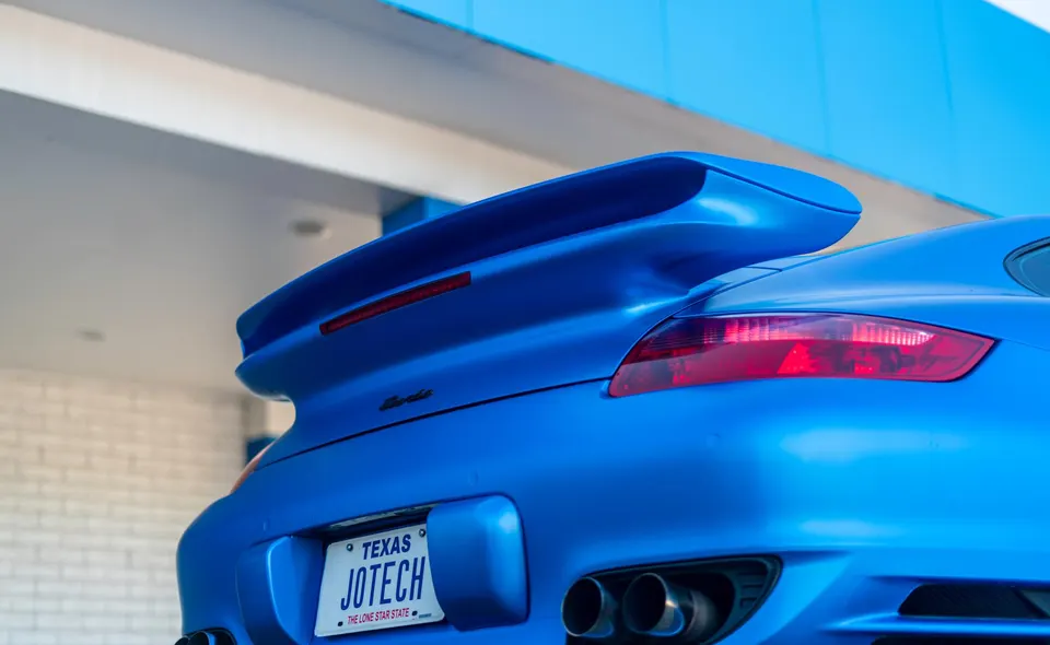 A close-up of the rear of a blue Porsche with a large rear wing, red taillights, and dual exhaust tips.