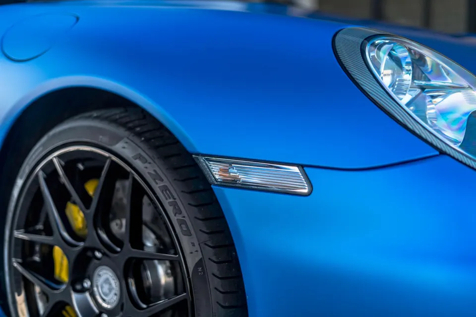 Close-up of a matte blue Porsche 911 front fender, black multi-spoke wheel, Pirelli P Zero tire, and yellow brake caliper.