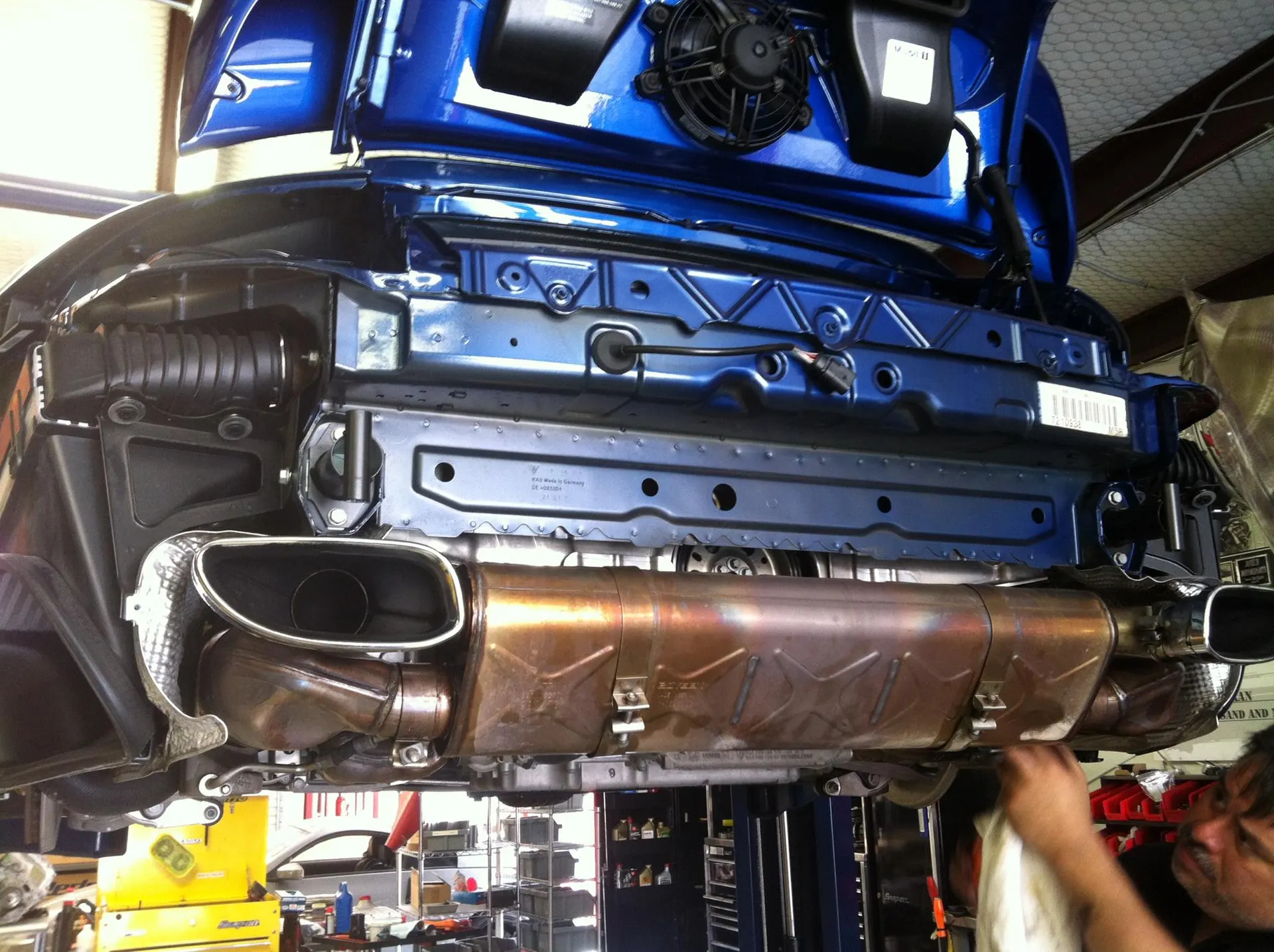 Underside view of a blue Porsche on a lift in a shop, showing the exhaust system and mufflers being worked on.