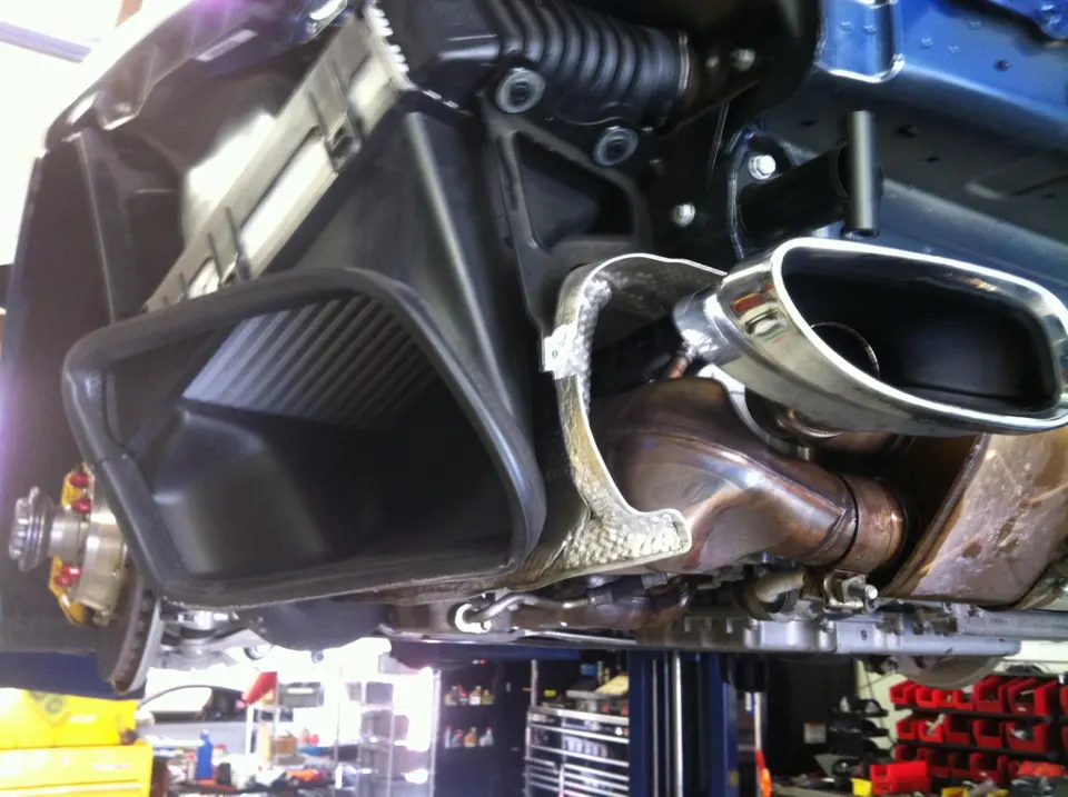 Underside view of a performance car on a lift, showcasing the exhaust system, muffler, and air intake duct in a shop.