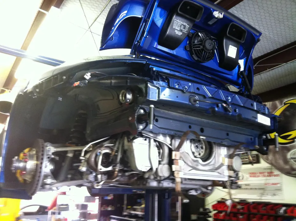 A blue Porsche 911 on a lift in a performance shop, with the rear bumper removed exposing the engine and turbochargers.