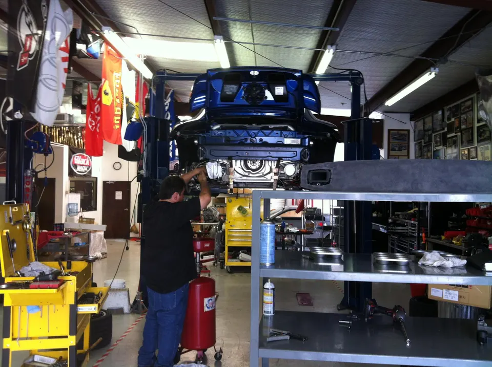 A mechanic works on the underside of a blue car lifted in a busy performance tuning shop.