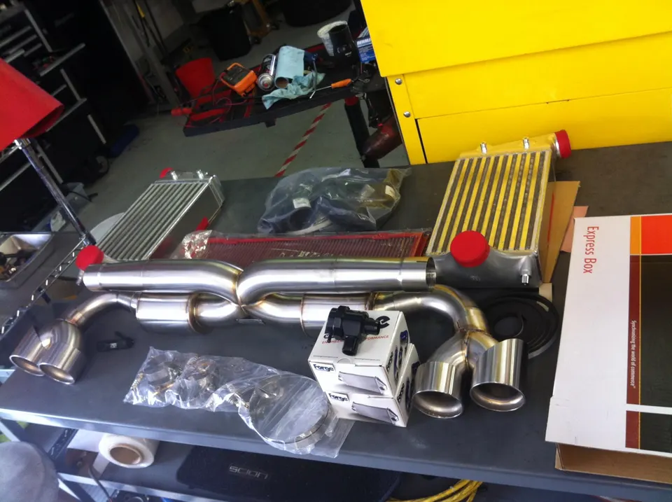 Performance car parts laid out on a table in a shop, including a stainless steel exhaust system, intercoolers, and a blow-off valve.