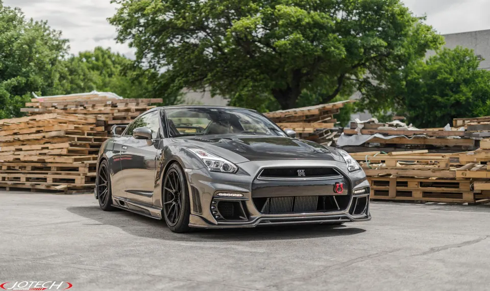 A grey modified Nissan GT-R with aftermarket wheels and a carbon fiber hood parked outdoors.