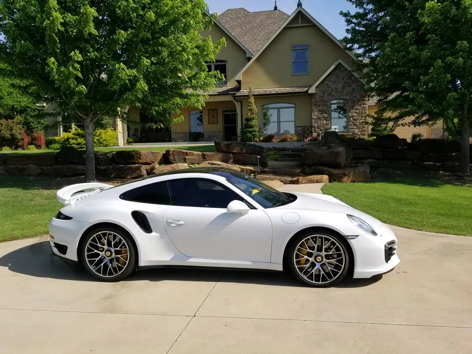 A white Porsche 911 Turbo S parked on a driveway in front of a large luxury home on a sunny day.
