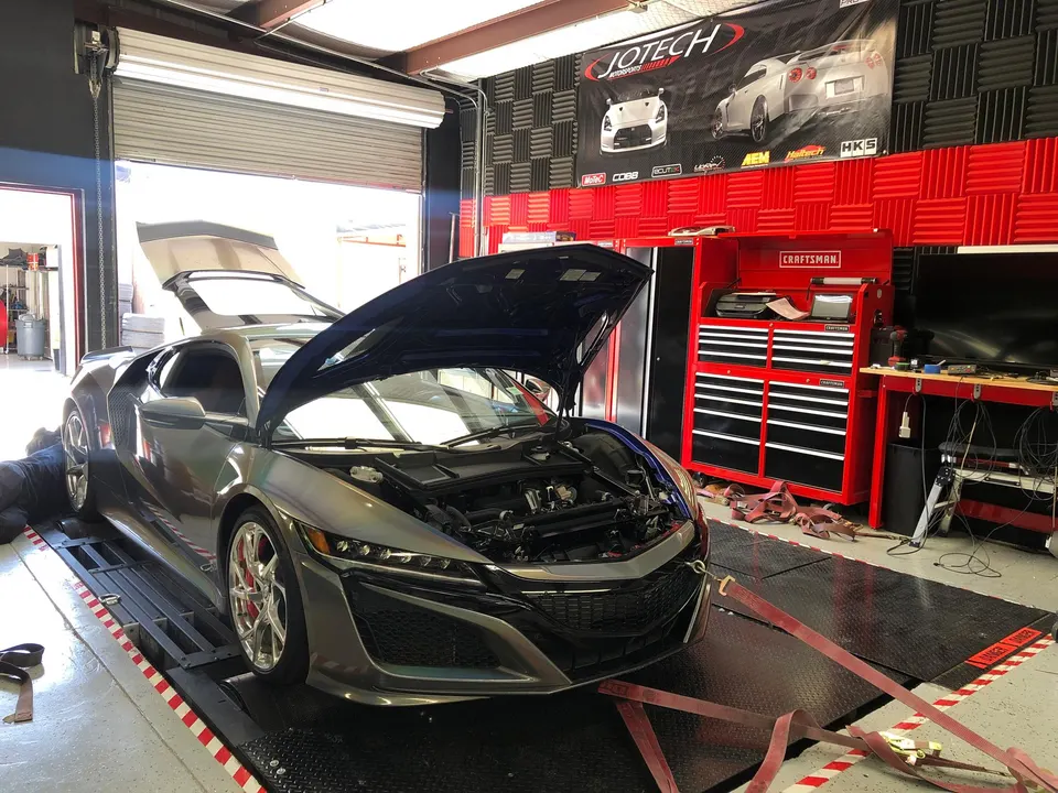A silver Acura NSX with its hood and engine cover open, strapped to a dyno in a performance tuning shop.