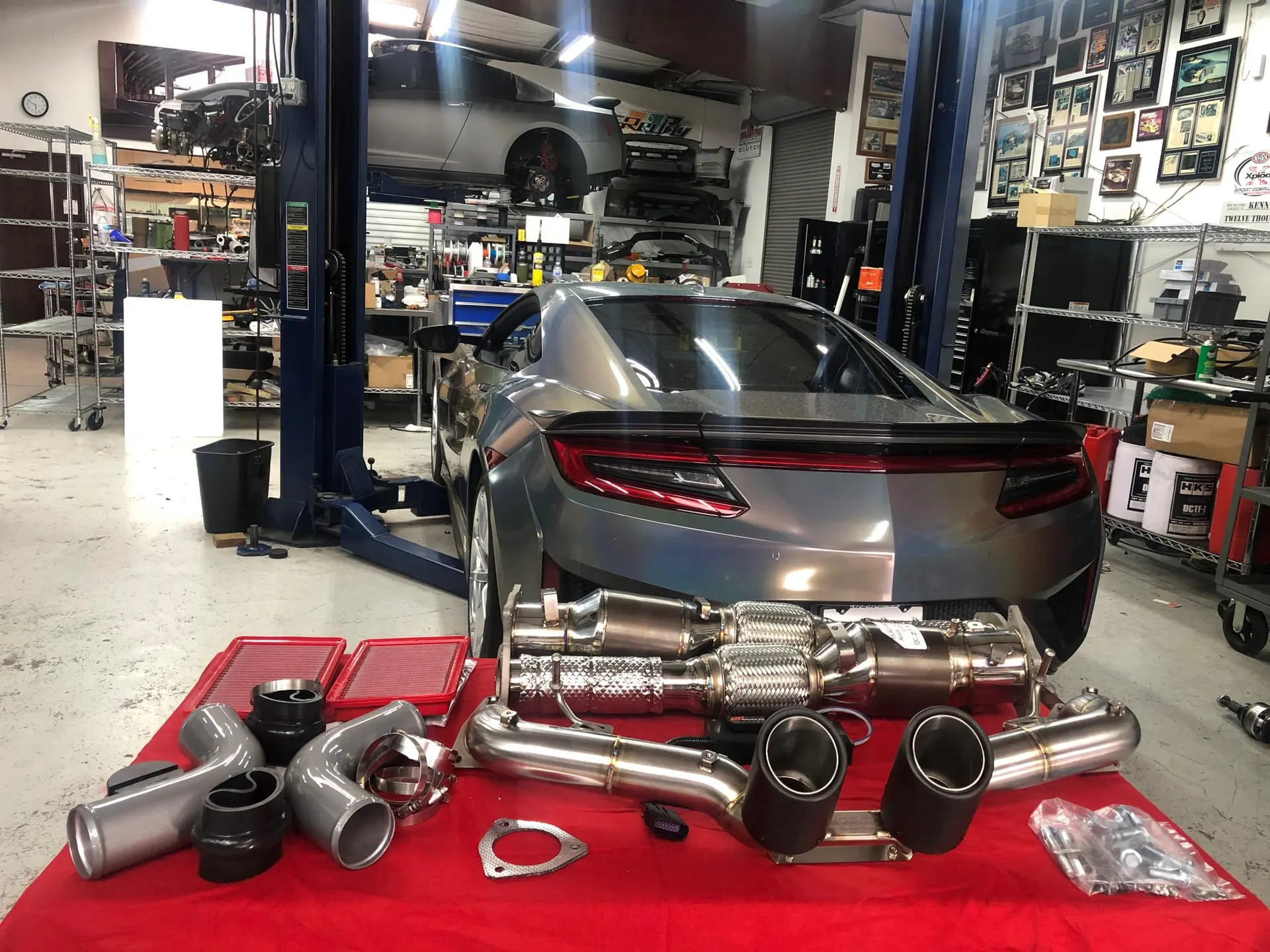 Acura NSX on a lift in a performance shop with an aftermarket exhaust system and intake parts laid out on a red cloth.